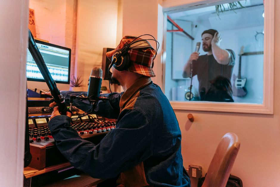Man using sound mixer in studio with vocalist in soundproof booth for recording session.